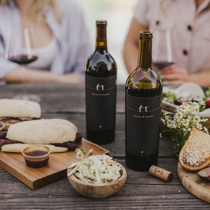Two open bottles of 2023 Harvey & Harriet Red Blend on a rustic table with plates of food surrounding it.