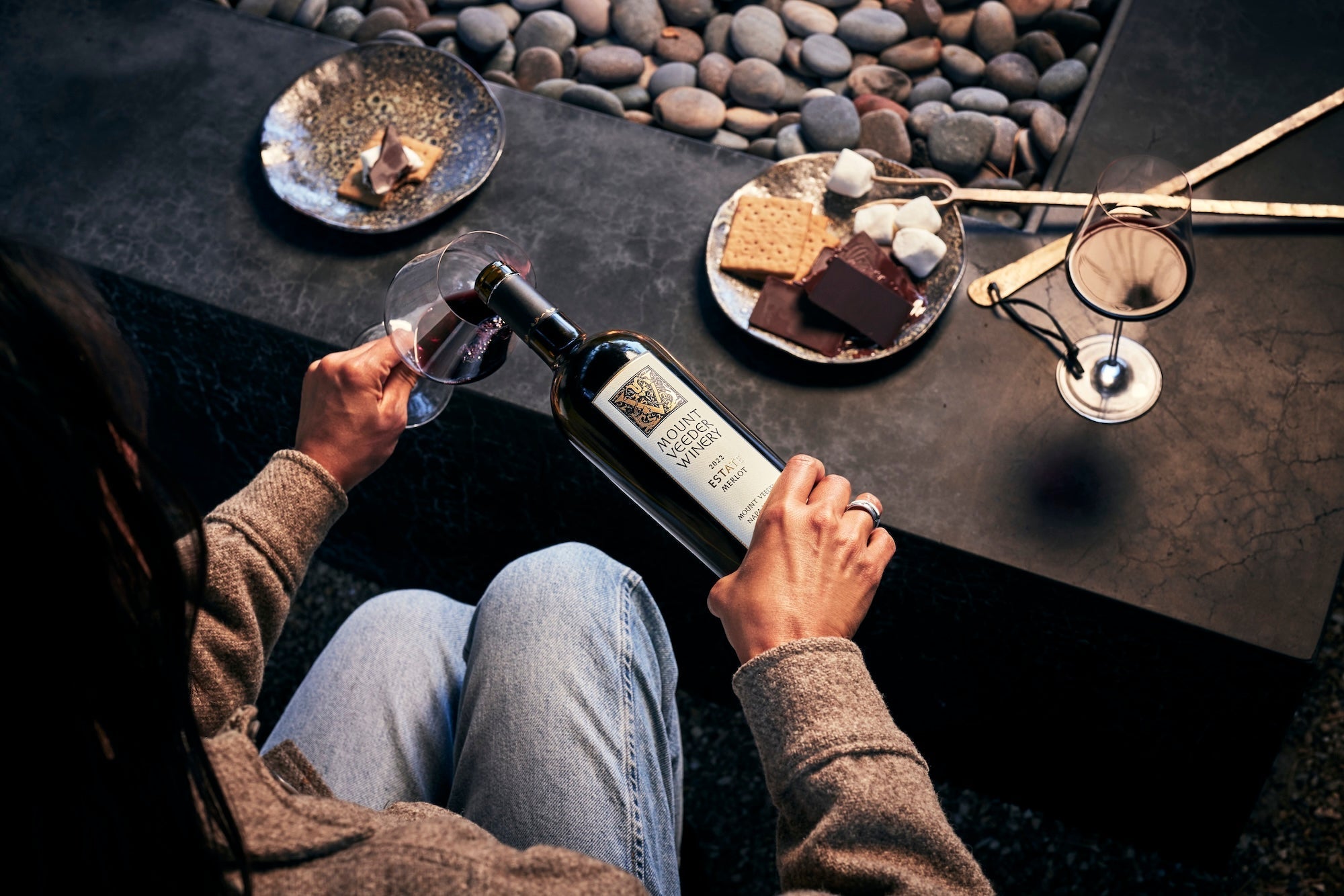 A woman in front of a fire pit pours the Estate Merlot into a glass with smores on the table 