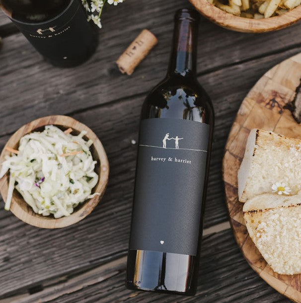 A bottle of 2023 Harvey & Harriet Red Blend laying label side up on a rustic table with plates of food surrounding it.