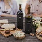 Two open bottles of 2023 Harvey & Harriet Red Blend on a rustic table with plates of food surrounding it.