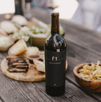 A bottle of 2023 Harvey & Harriet Red Blend on a rustic table with plates of food surrounding it.