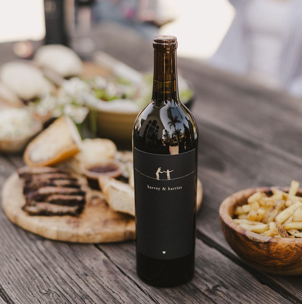 A bottle of 2023 Harvey & Harriet Red Blend on a rustic table with plates of food surrounding it.