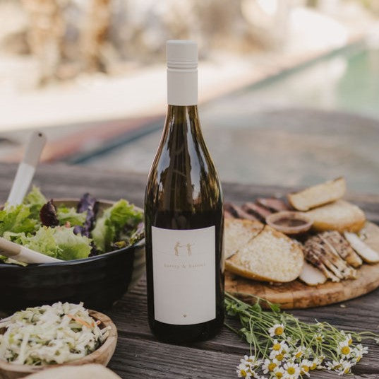 A bottle of 2021 Harvey & Harriet White Blend on a rustic table with plates of food surrounding it.