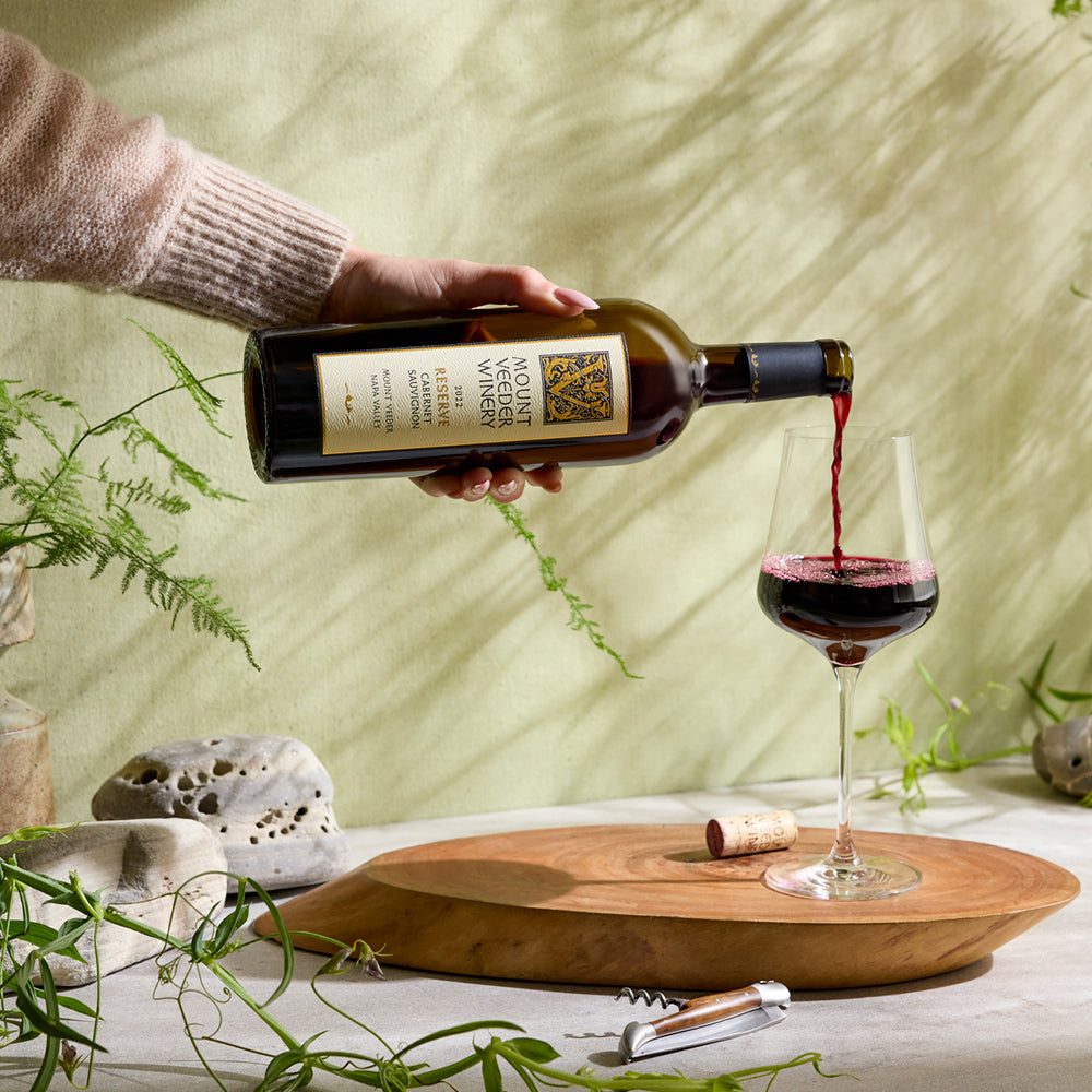 A person pours a bottle of Mount Veeder Reserve Cabernet Sauvignon into a glass on a wooden surface