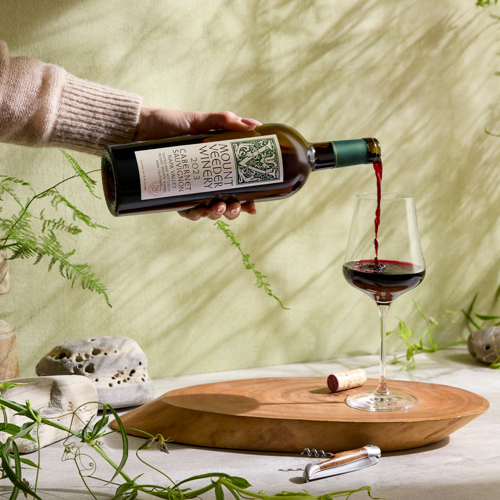 A person pours a bottle of Mount Veeder Reserve Cabernet Sauvignon into a glass on a wooden surface