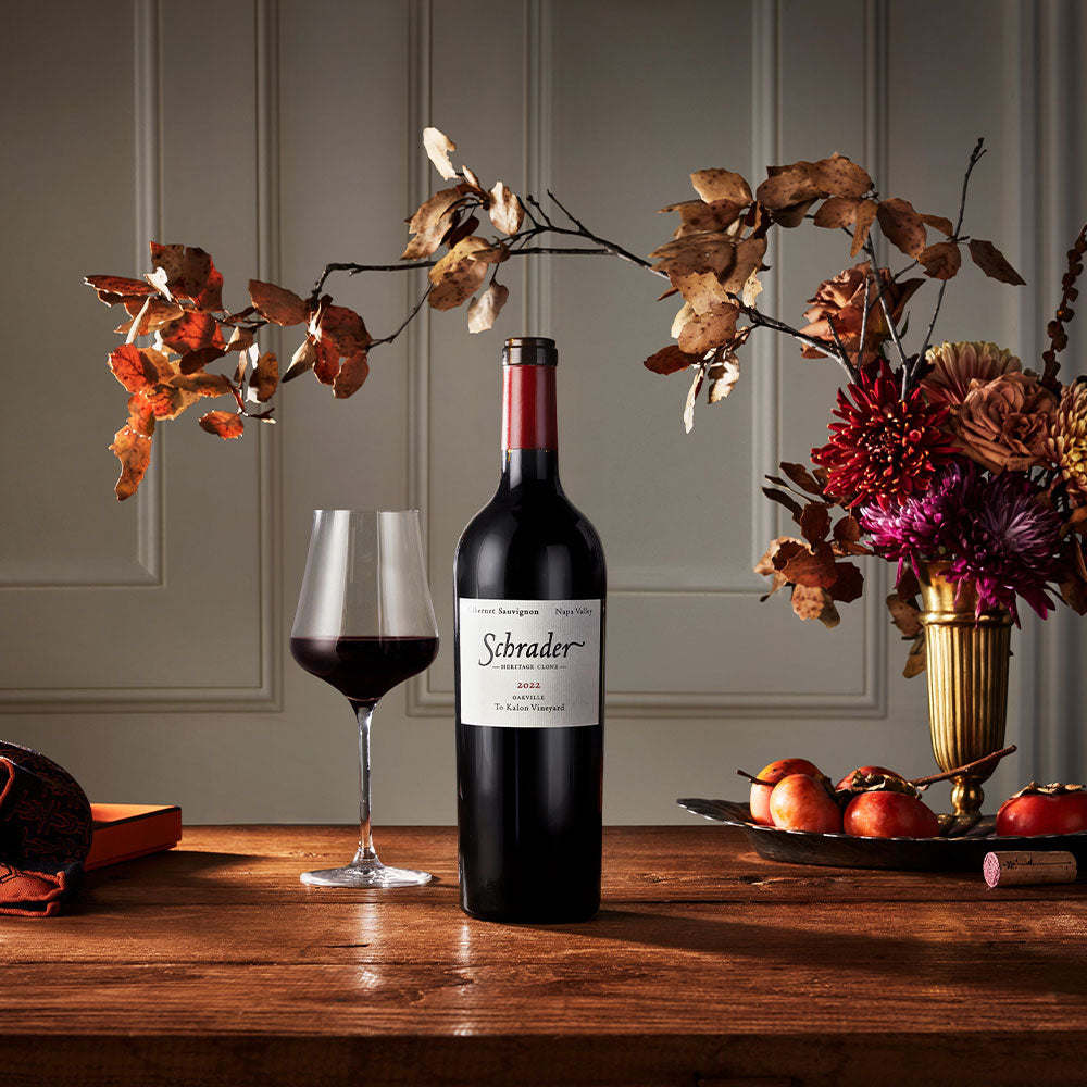 A bottle of Schrader 2023 red wine stands on a wooden table beside a filled wine glass, with autumn flowers and apples arranged around it in a warm, elegant interior setting.