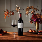 A bottle of Schrader 2023 red wine stands on a wooden table beside a filled wine glass, with autumn flowers and apples arranged around it in a warm, elegant interior setting.