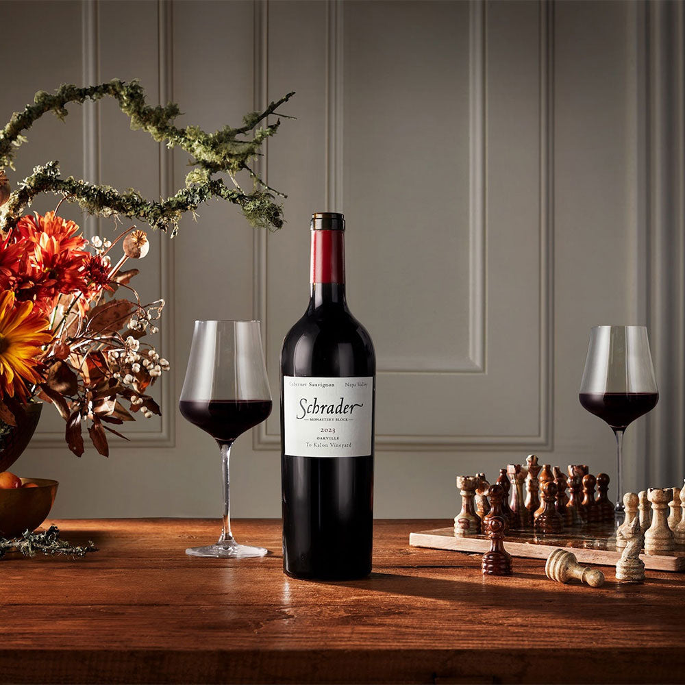 A bottle of Schrader 20123 Monastery Block Cabernet Sauvignon stands on a wooden table between two filled wine glasses, with decorative flowers on the left and a chess set on the right against a paneled gray wall.
