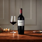 A bottle of Schrader 2023 WH Cabernet Sauvignon stands on a wooden table beside a filled wine glass, with a softly lit interior wall in the background.